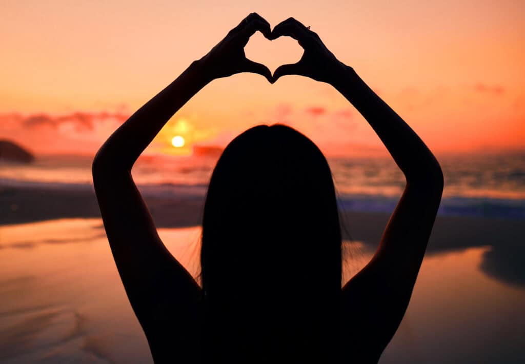 Sunset with a woman's silhouette and arms overhead with hands touching so they form a heart in the center.